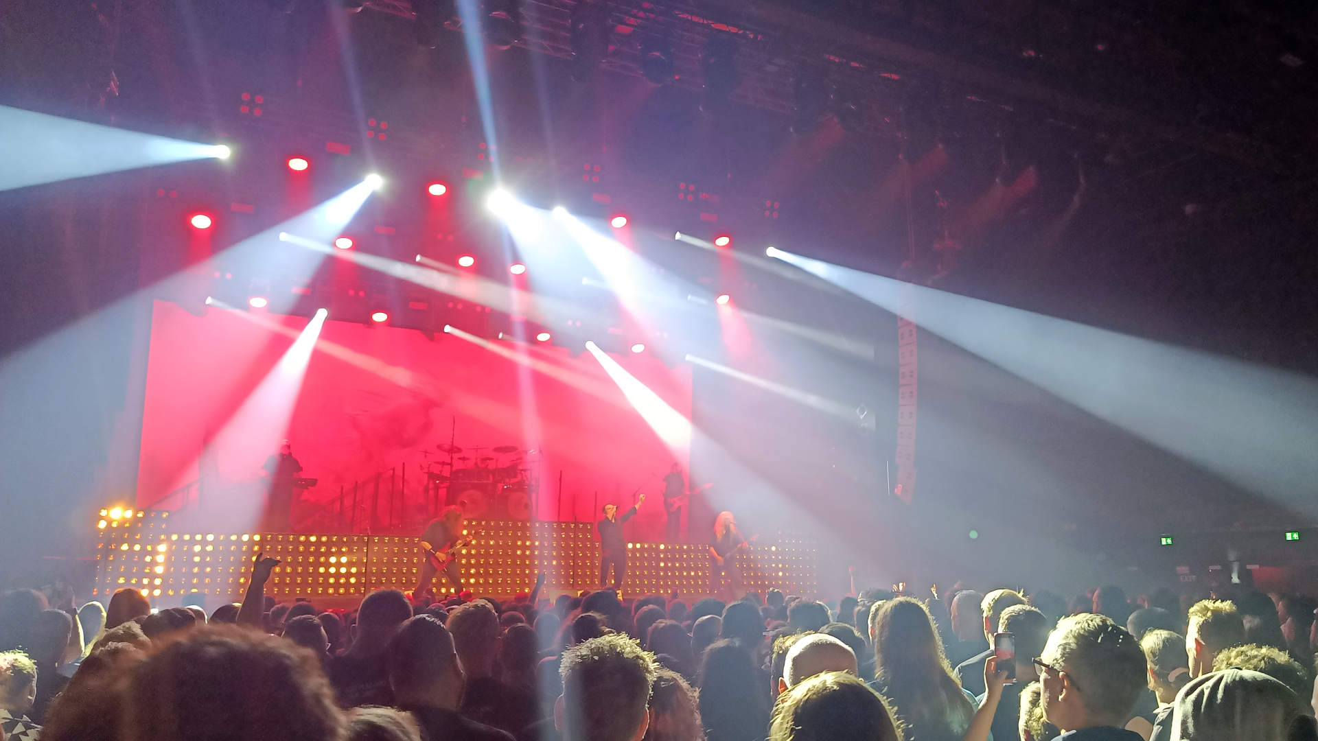 Concertfoto: band op podium met rood achtergrondlicht en spotlights die op de zaal schijnen